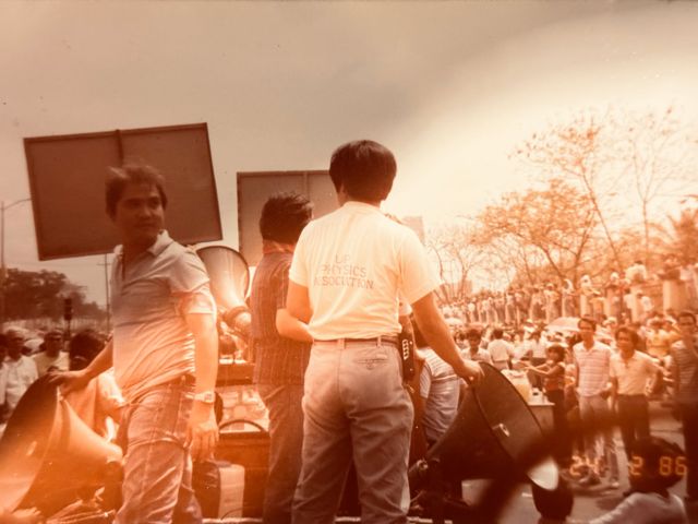 Faded color photograph dated February 24, 1986, of men on a truck with megaphones above a large street crowd, one wearing a shirt printed UP Physics Association
