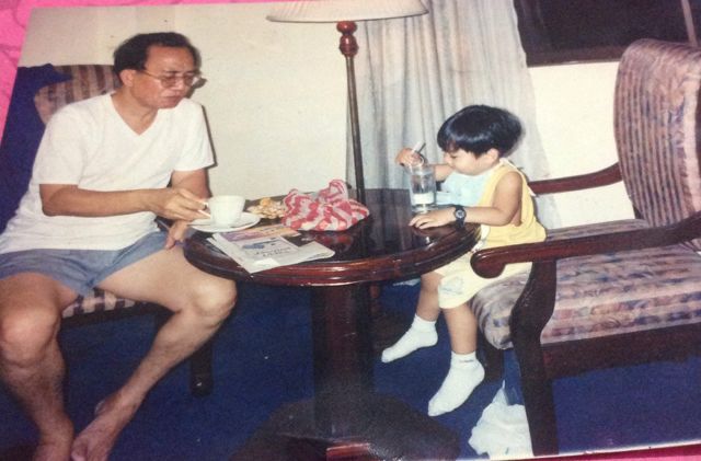 Dad in a white shirt having coffee at a small round table, with Gregor as a toddler beside him stirring a glass of milk
