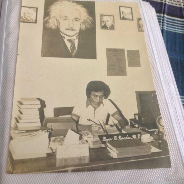 Dad at his office desk writing, under a large portrait of Albert Einstein and smaller framed portraits of other physicists, with his nameplate on the desk