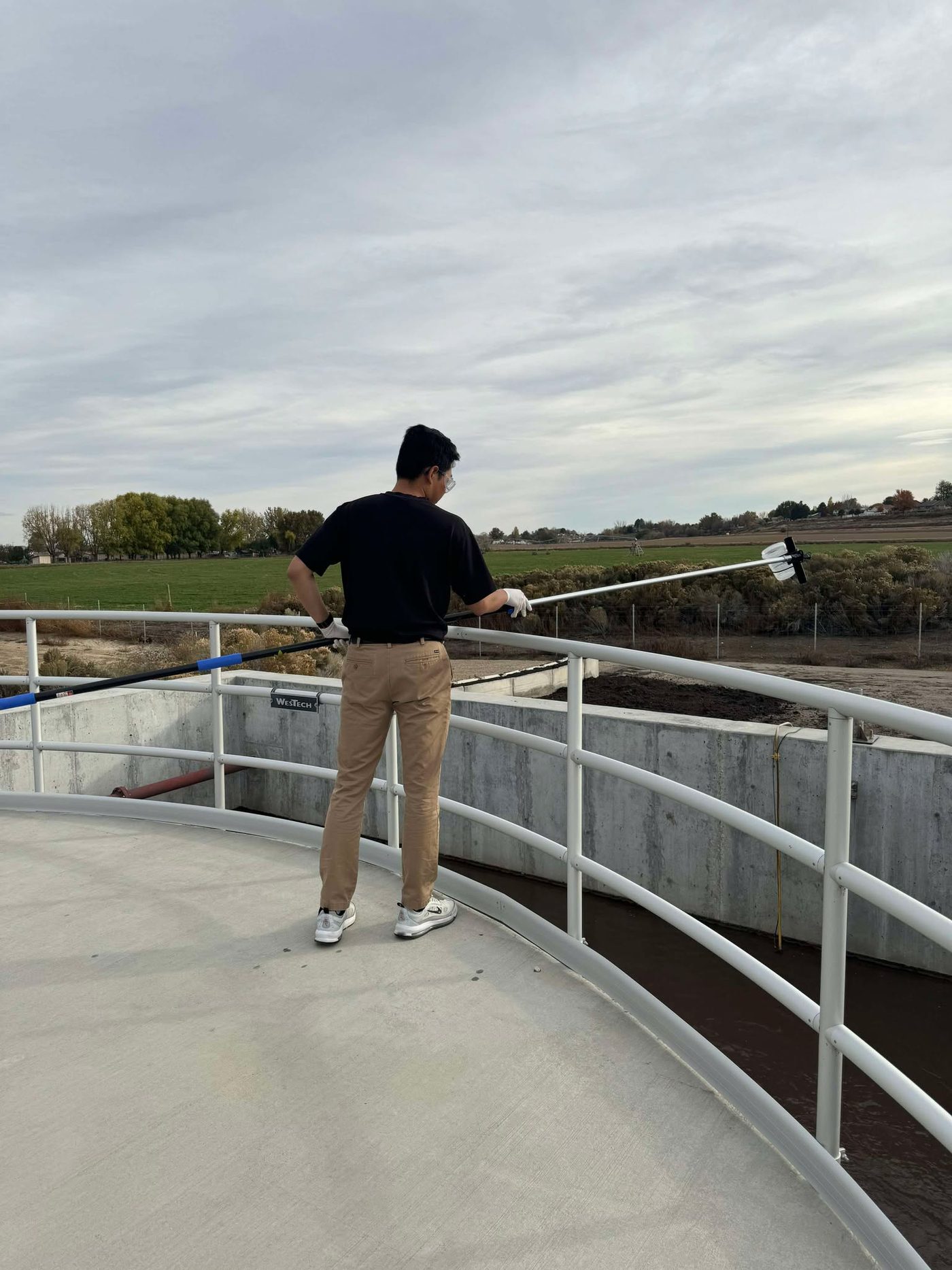 Gregor on the walkway of an oxidation ditch, reaching out over the railing with a long sampling pole to collect activated sludge