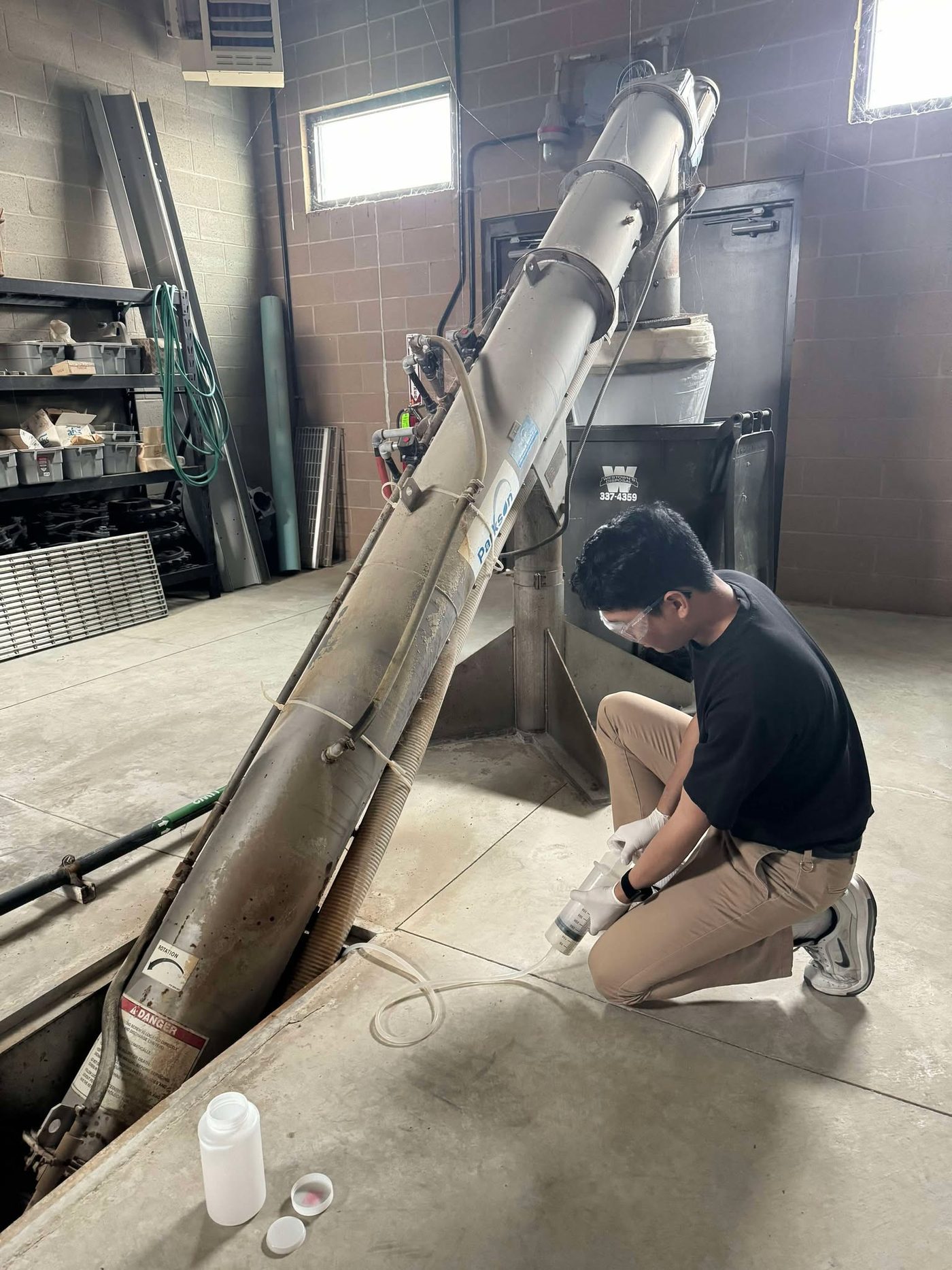 Gregor crouching beside an inclined screw conveyor at the plant headworks, drawing a sample with a syringe