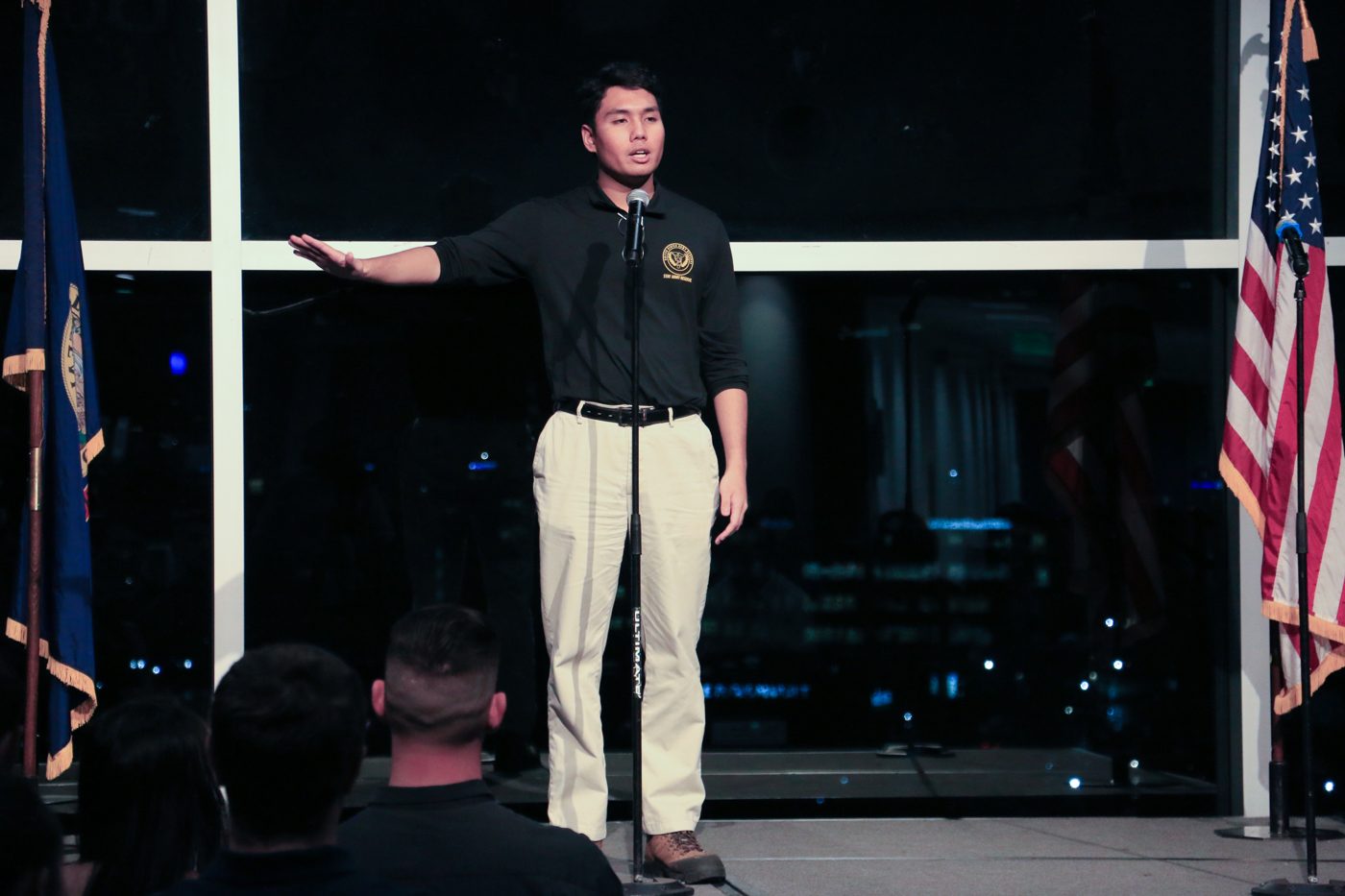Gregor at the microphone in a black Army Reserve polo, one arm extended mid-story, an Idaho flag and an American flag on either side