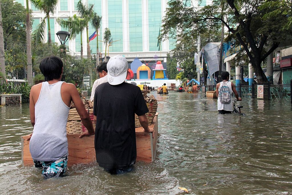 Two men push a wooden handcart through brown floodwater along a city street; more people wade behind them.