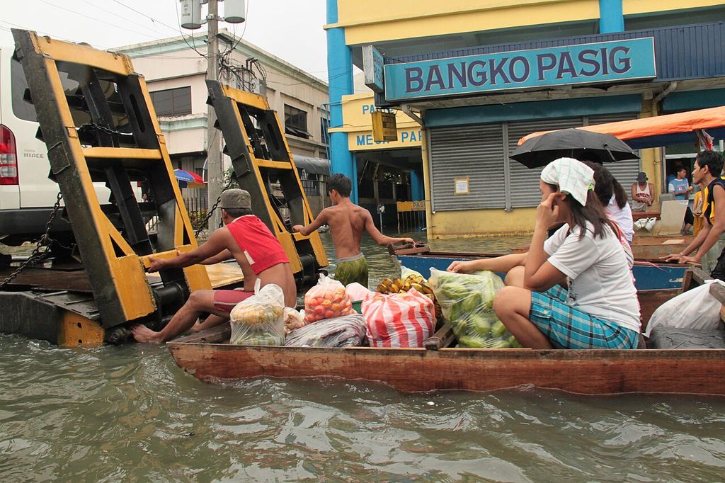 A wooden boat loaded with sacks of vegetables and passengers moves along a flooded street past a shop sign reading Bangko Pasig.