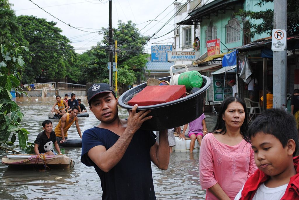 A man carries a basin of belongings on his shoulder through floodwater, followed by a woman and a boy; others float on rafts behind.
