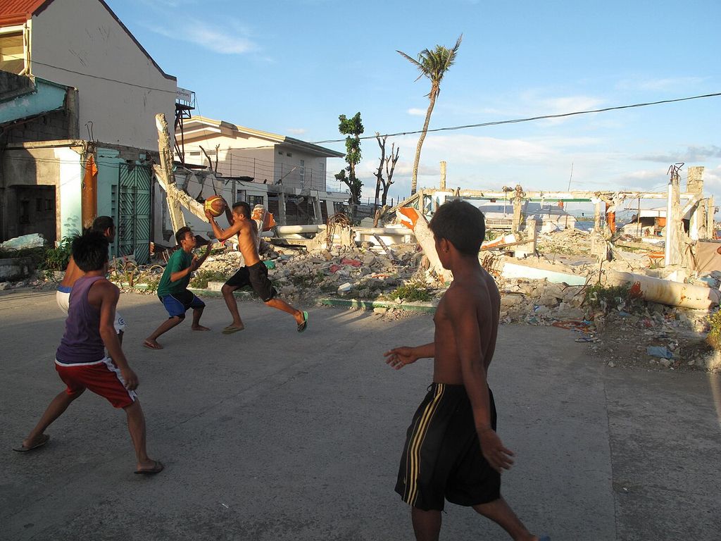 Boys play basketball on a cleared court, with storm debris and damaged buildings behind them.