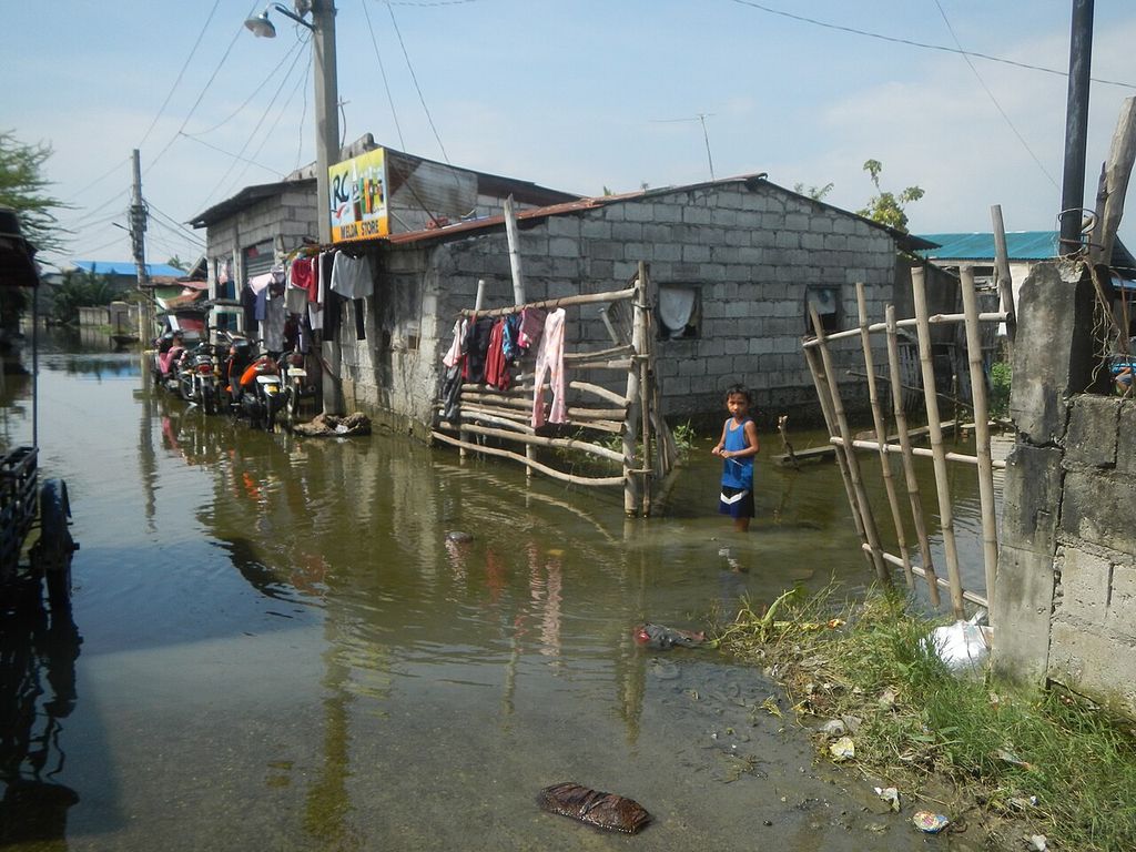 Floodwater stands around concrete-block houses with laundry hanging out; a child stands in the water.