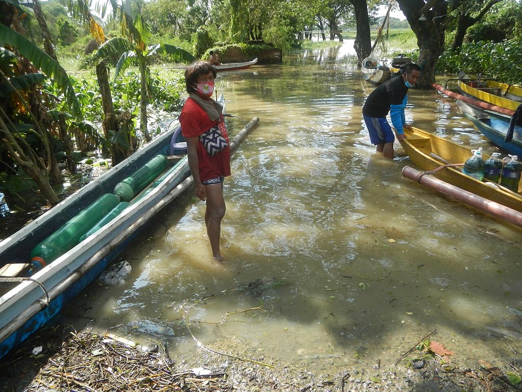 A man stands knee-deep in floodwater beside narrow wooden boats; another person wades nearby among banana trees.