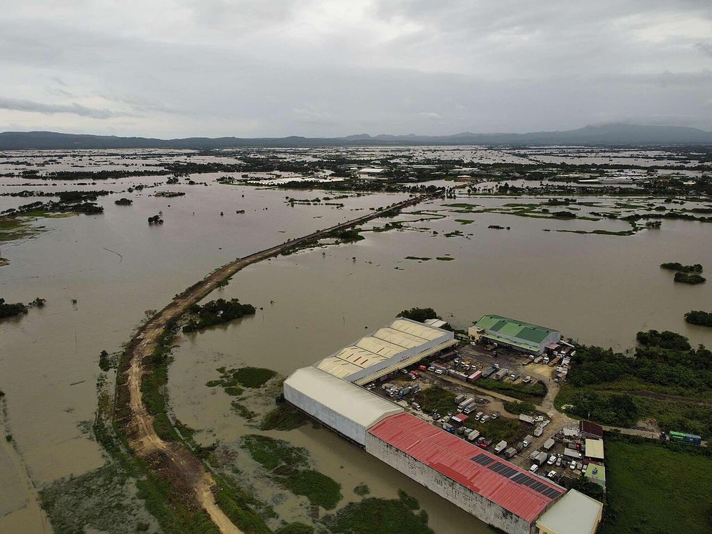 Aerial view of rice fields under brown floodwater stretching to the horizon, crossed by a raised road.