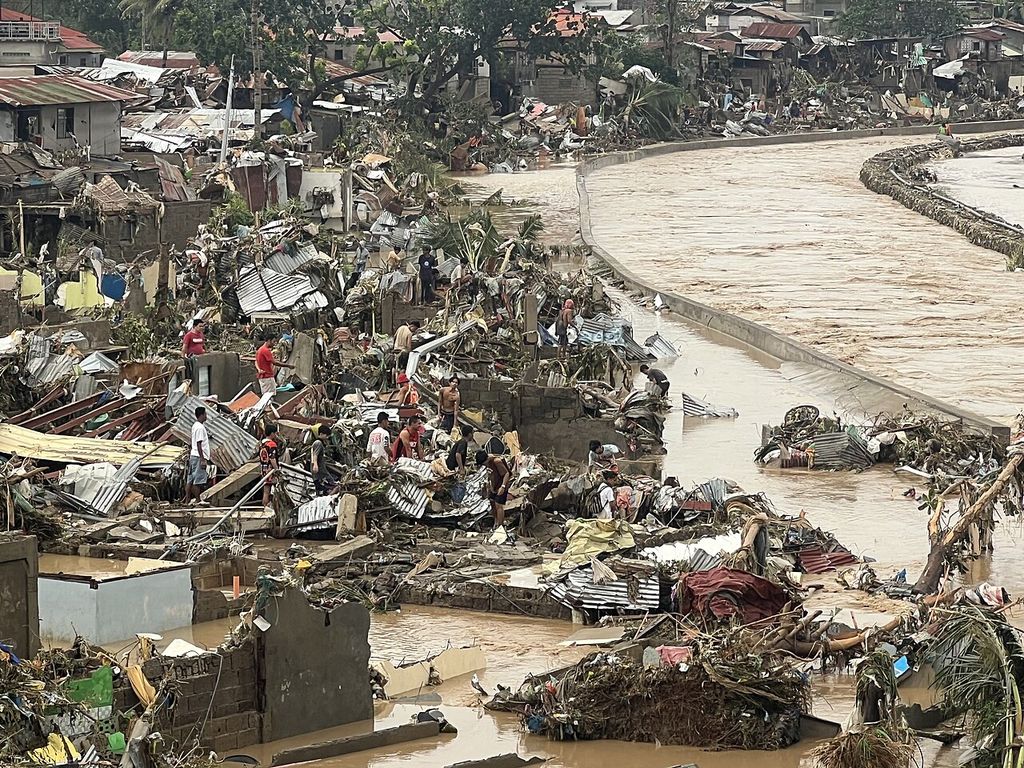 The swollen brown Mananga River beside a bank of destroyed houses and debris, with people among the wreckage.